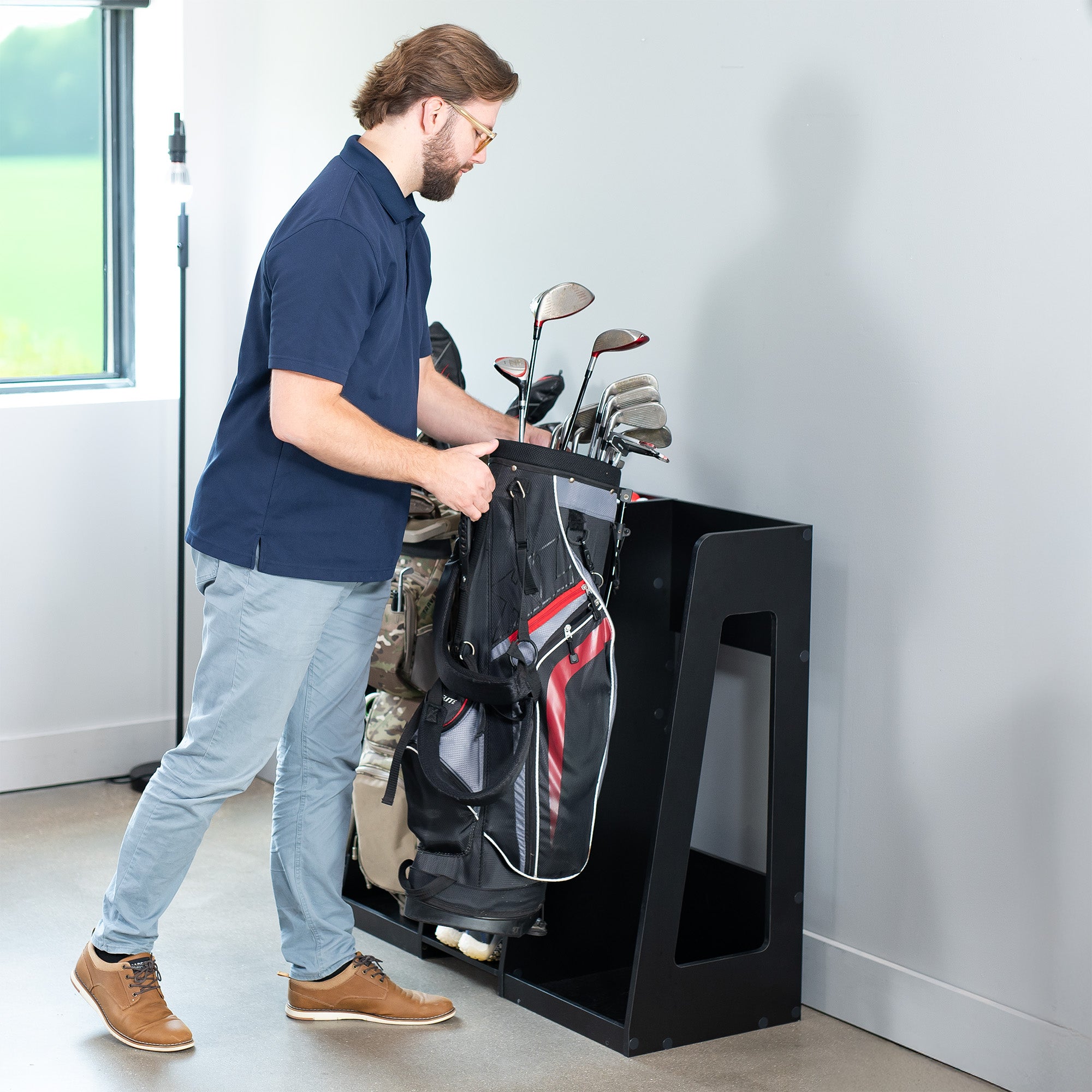 Man loading golf clubs into a black golf bag stand indoors.