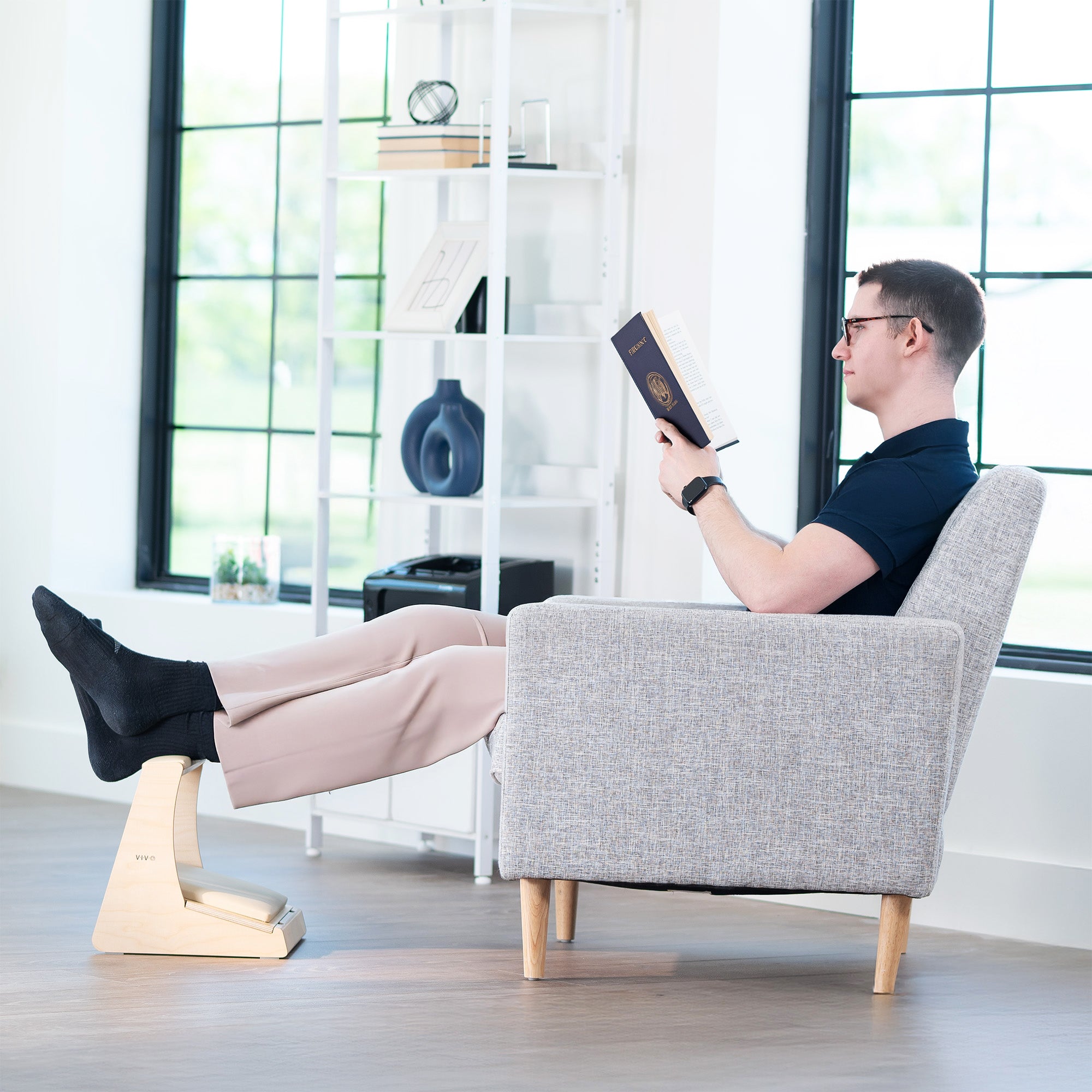 Man reading a book in a gray armchair with a footstool in a bright room.