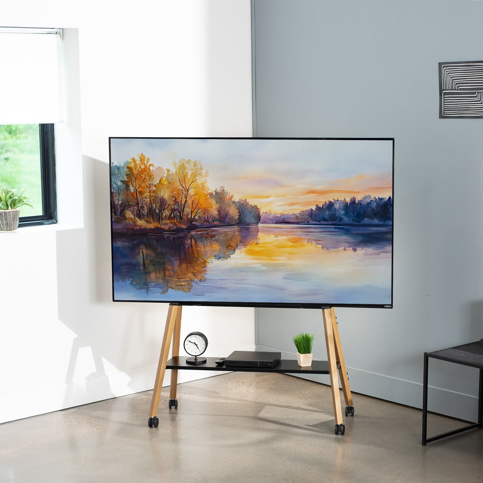 A large flat-screen TV on a wooden rolling stand in a modern room. The TV displays a colorful landscape painting of a lake at sunset.