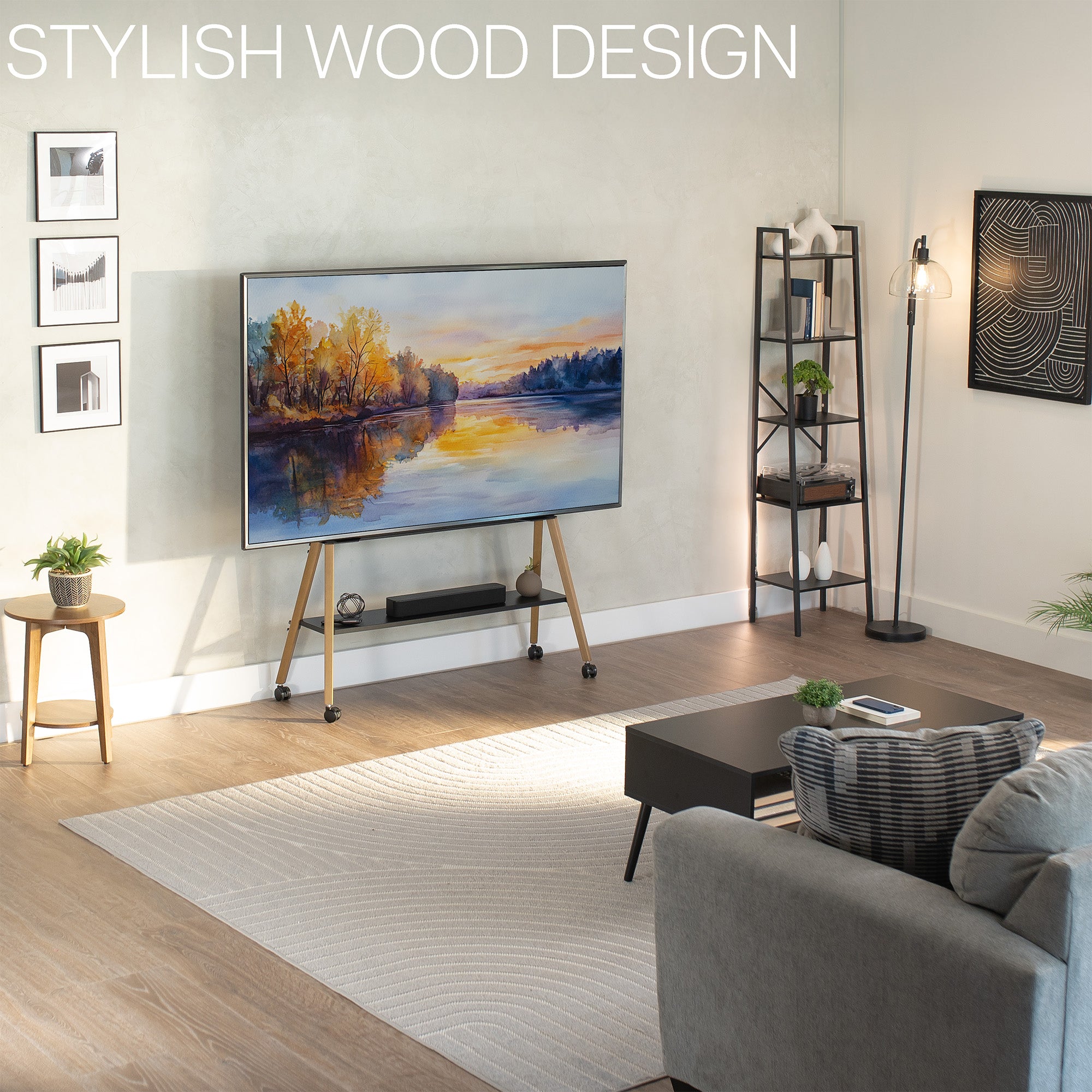 A modern living room scene showing a large wall-mounted TV on a slim wooden rolling stand with casters. The room has light wood flooring, a cream rug, a gray sofa, a black coffee table, a floor lamp, and framed wall art.