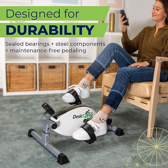 A woman sits on a wooden chair in a home office setting, using a white and black desk exercise bike. The image features promotional text overlays highlighting the bike's durability and features.