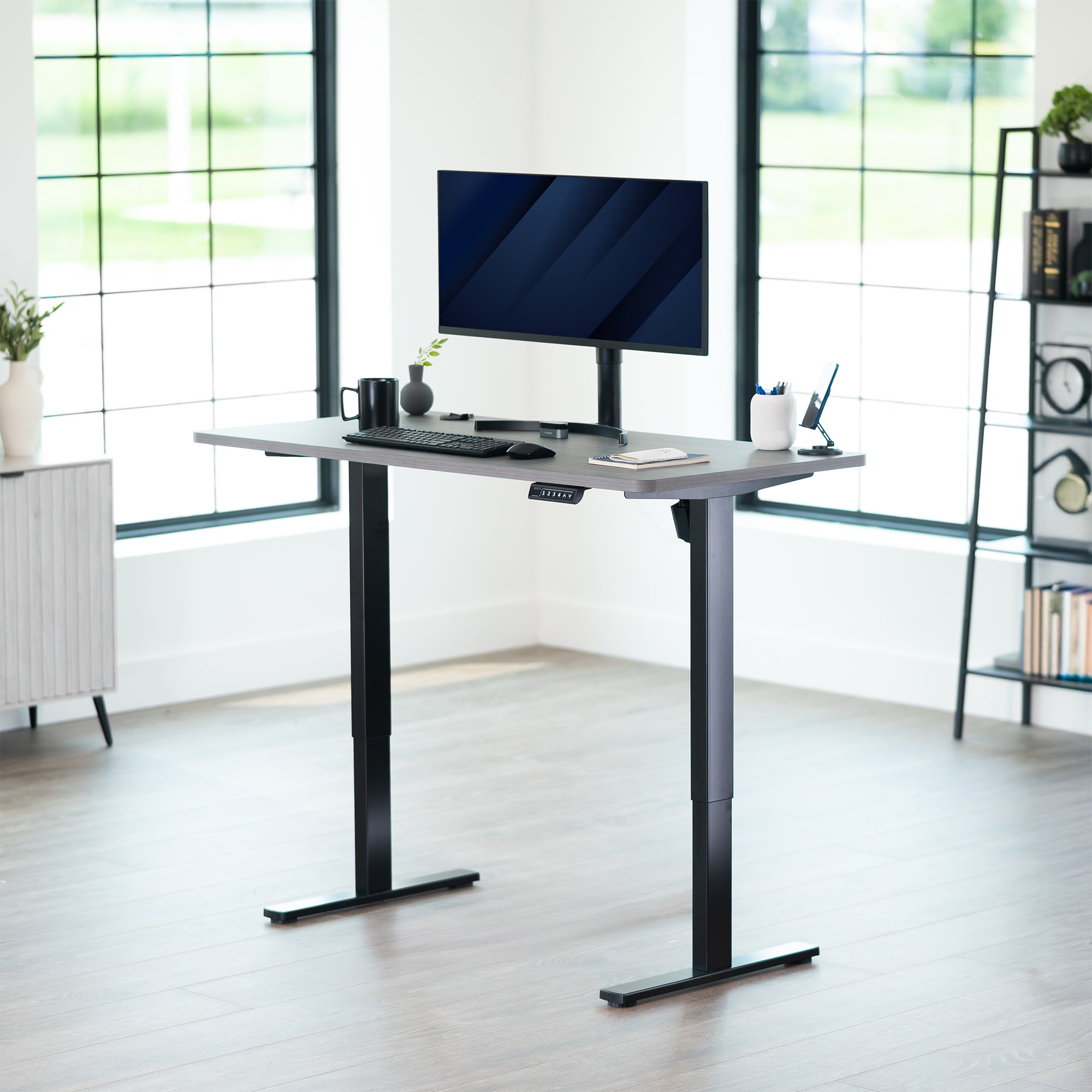 Adjustable height desk with computer setup in a bright room with large windows.