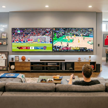 A man sits on a gray sectional sofa in a modern living room, watching two large wall-mounted TVs showing sports broadcasts. The room includes a wooden media console, coffee table, and framed wall art.