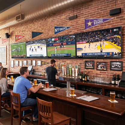 A cozy sports bar interior with a wooden bar counter, brick wall, and multiple television screens showing basketball and baseball games. Several people are seated at the bar, and sports pennants hang on the wall.