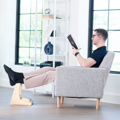 Man reading a book in a gray armchair with a footstool in a bright room.