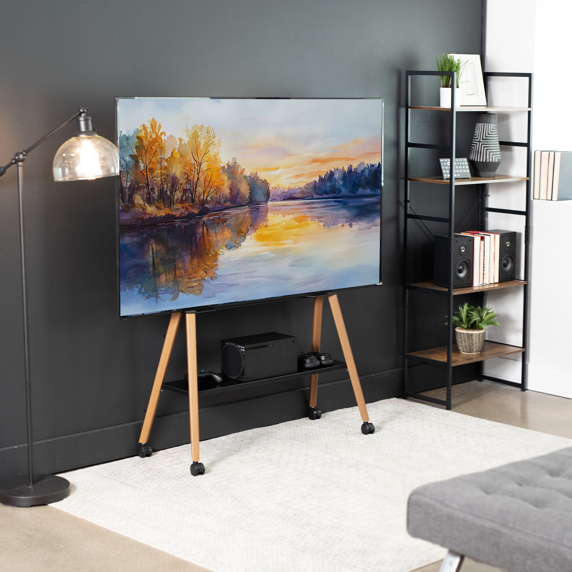 A large flat-screen TV on a black metal and walnut wood easel stand in a modern living room setting. The TV displays a colorful landscape painting of a lake at sunset.