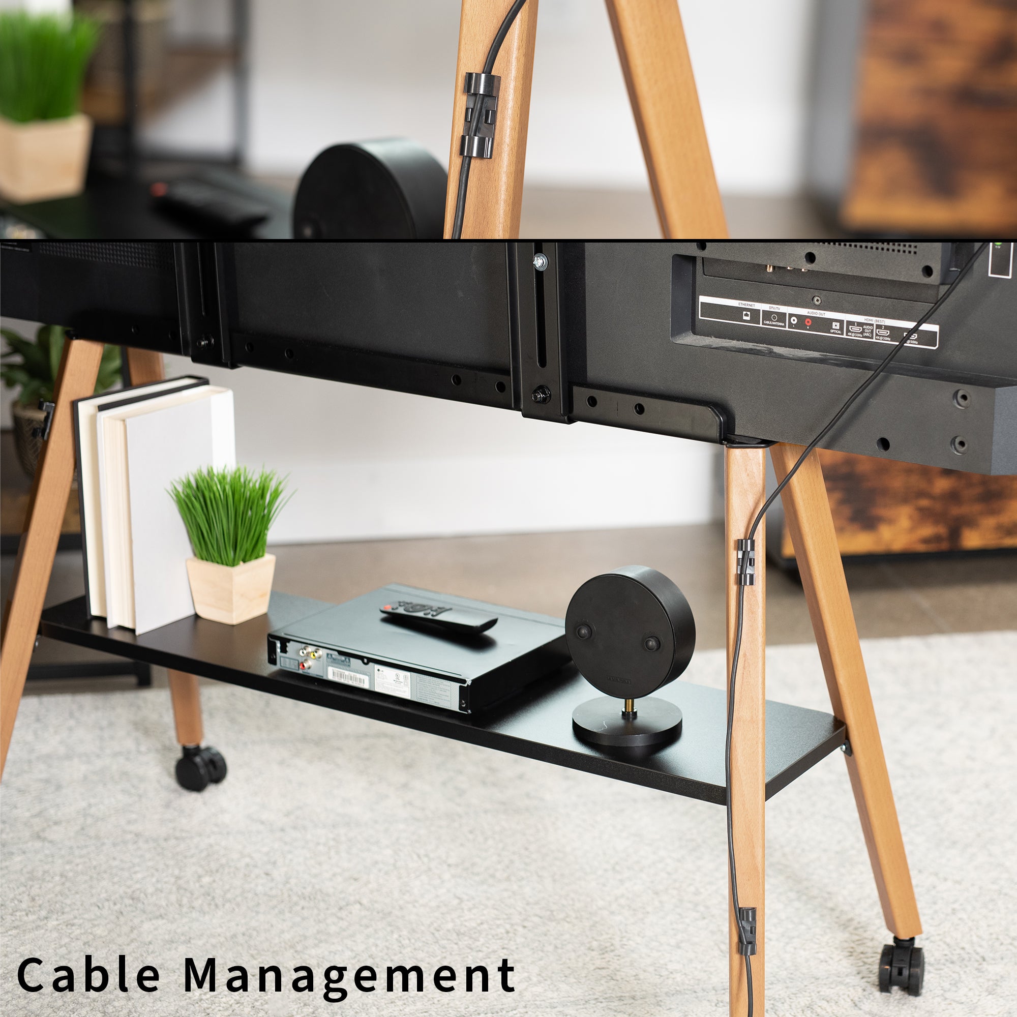 A close-up of a black cable management clip mounted on a wooden rolling TV stand in a living room setting. The lower shelf of the stand holds a cable modem, a smart speaker, and a small potted plant, with a TV mounted above.