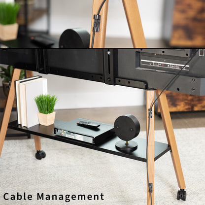 A close-up of a black cable management clip mounted on a wooden rolling TV stand in a living room setting. The lower shelf of the stand holds a cable modem, a smart speaker, and a small potted plant, with a TV mounted above.