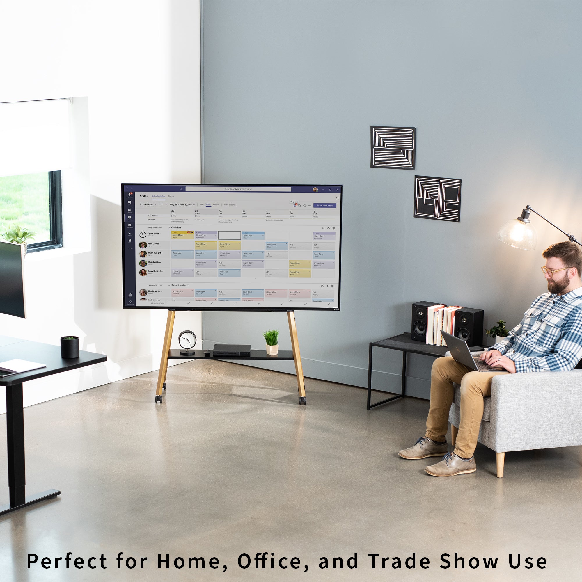A large TV screen on a wooden stand in a modern office space, with a man sitting in a gray armchair to the right working on a laptop. The screen displays a colorful meeting agenda or calendar interface.