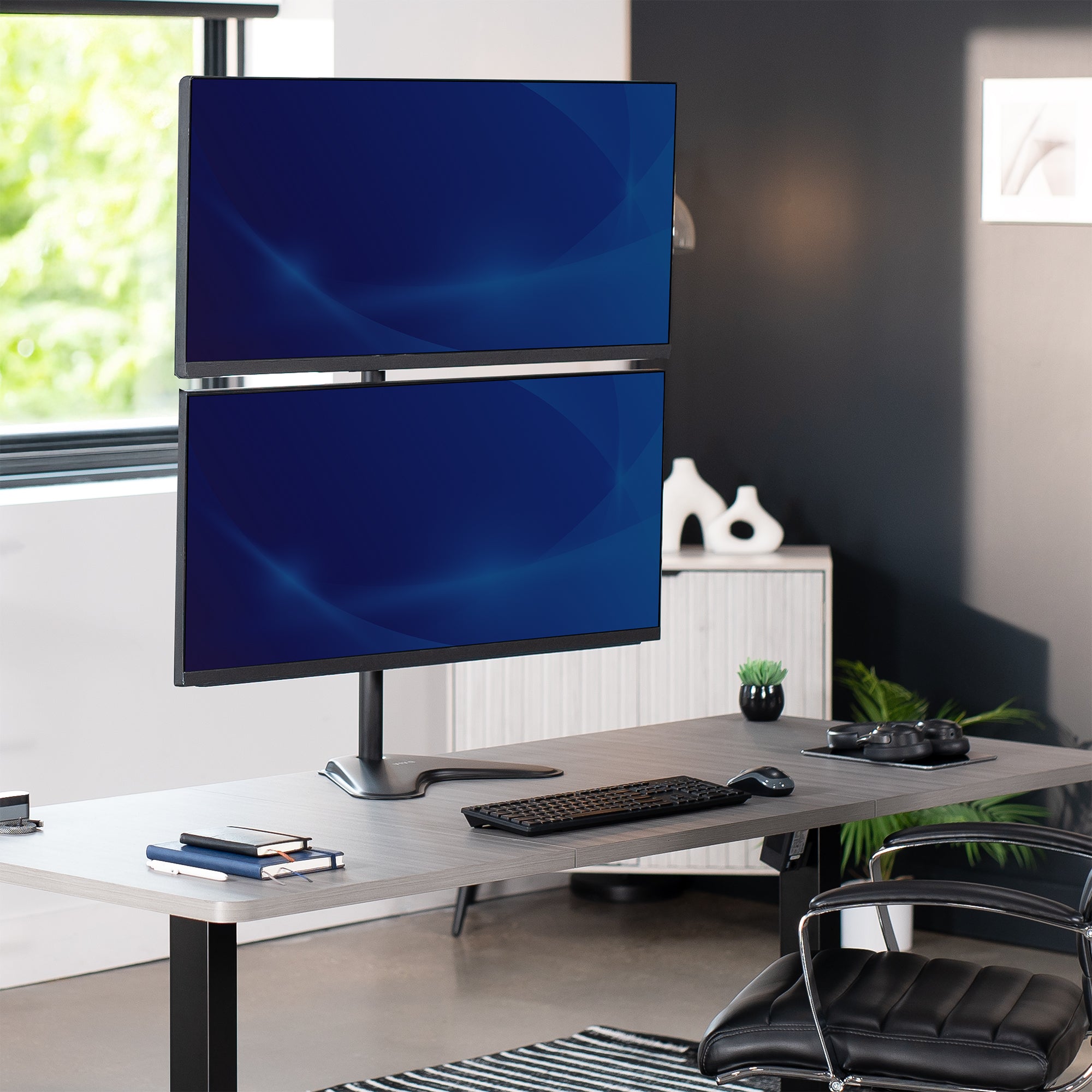 Two computer monitors on a freestanding monitor stand on a desk in an office setting with a chair and decor.