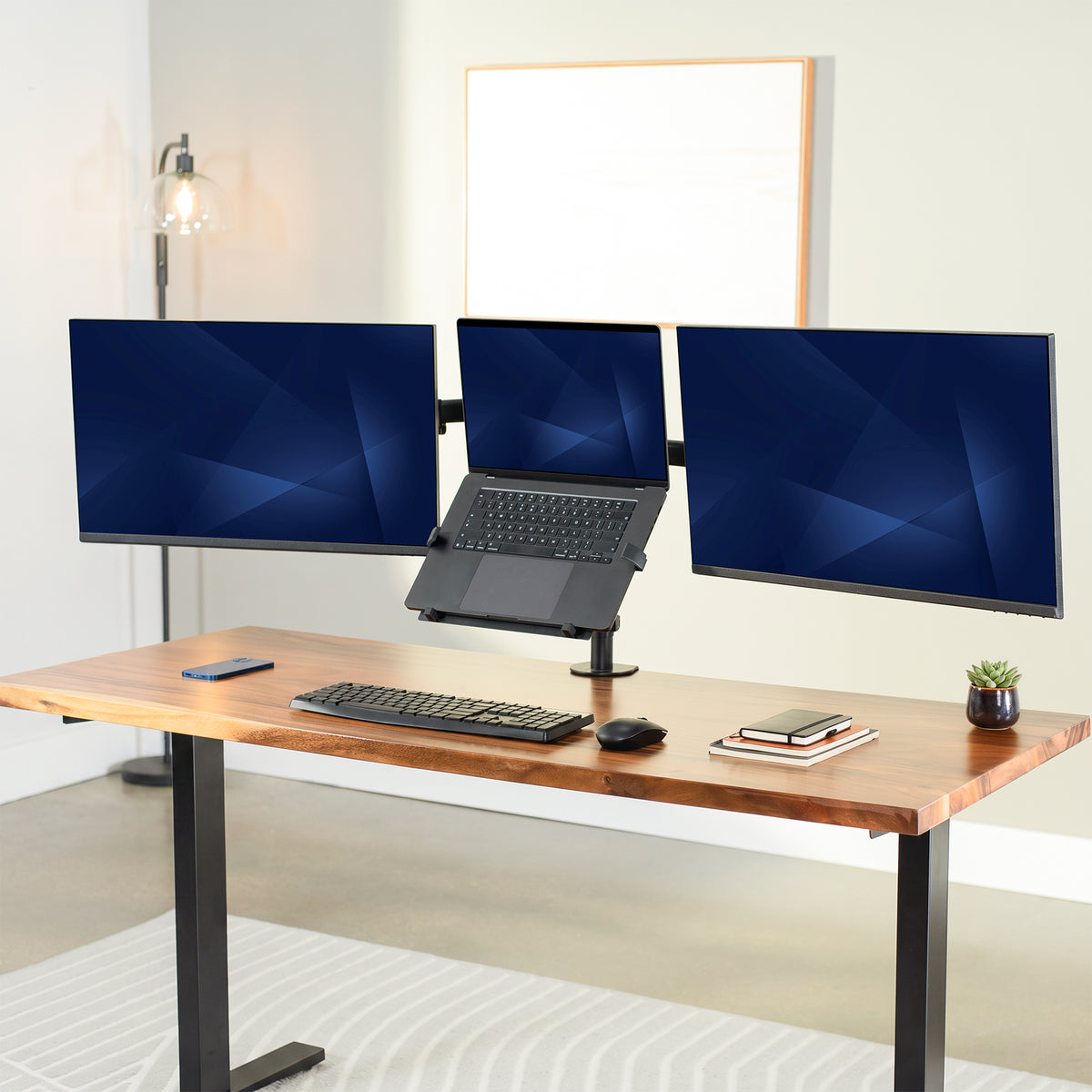 A modern standing desk setup with a wooden desktop and black metal legs, featuring a triple-monitor desk stand with a laptop in the center, alongside a keyboard, mouse, phone, and small plant.