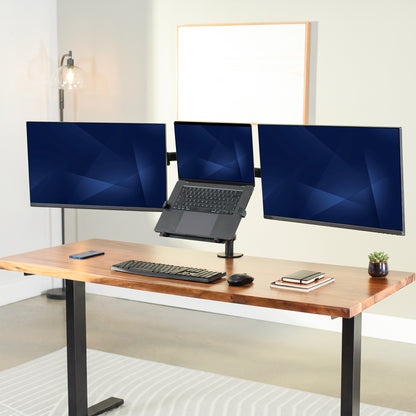 A modern standing desk setup with a wooden desktop and black metal legs, featuring a triple-monitor desk stand with a laptop in the center, alongside a keyboard, mouse, phone, and small plant.