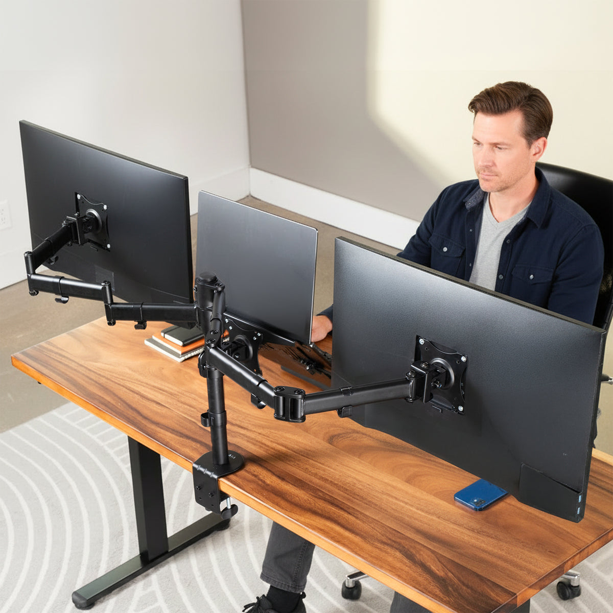 A man sits at a wooden standing desk using dual monitor arm setups with a laptop in between computer monitors and a smartphone in a modern home office setting.