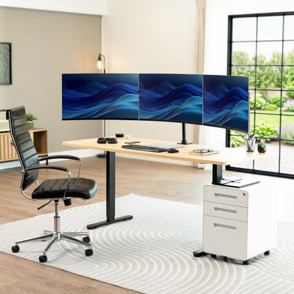 A modern home office setup featuring a light wood standing desk with a black metal frame, a curved triple-monitor desktop setup, a black leather office chair, a white rolling filing cabinet, and a gray patterned rug in a bright room with a large window.