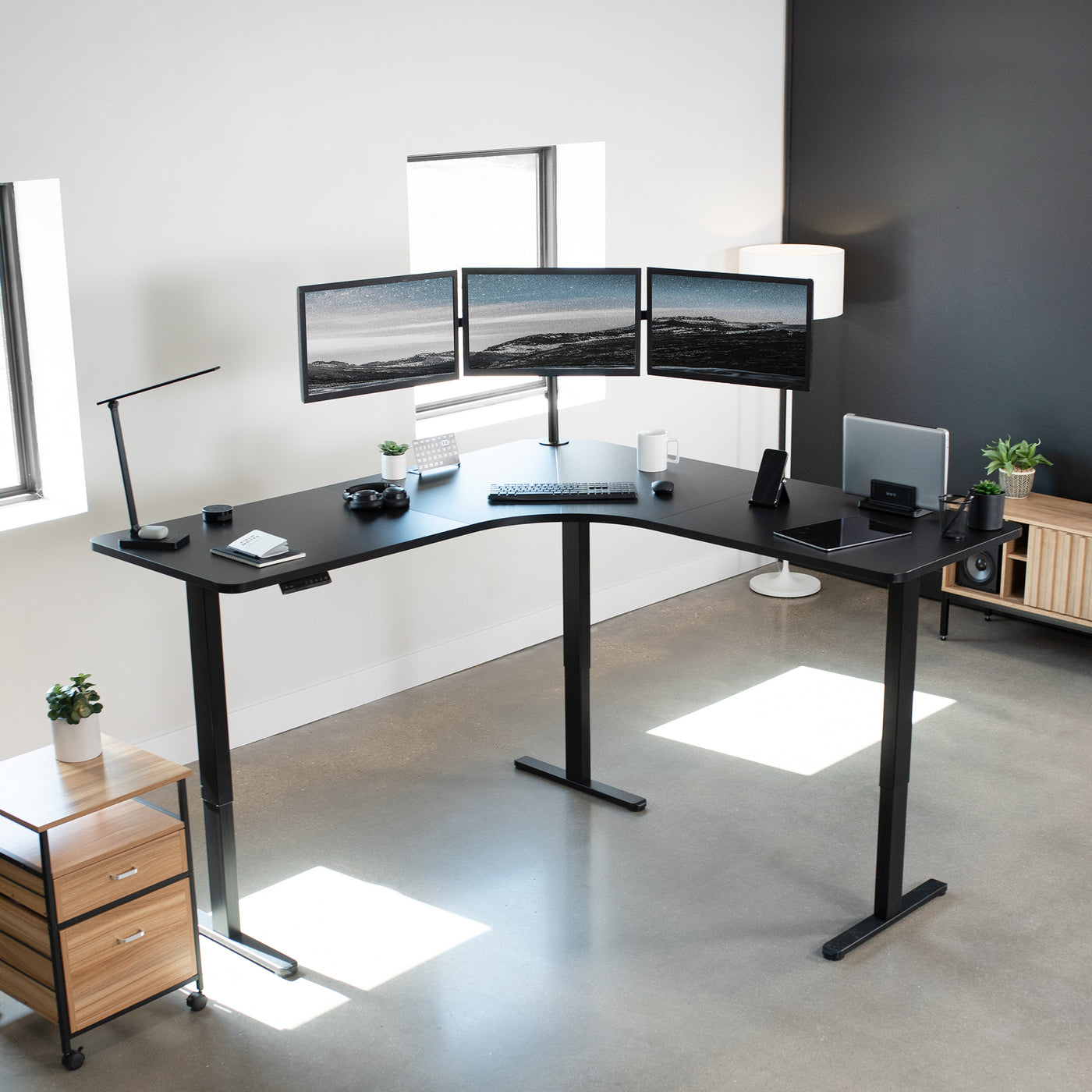 Standing desk in modern office space by windows with three monitors on the tabletop.