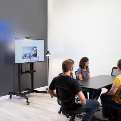 Electric height adjustable TV stand on a rolling caster wheel in a conference room.